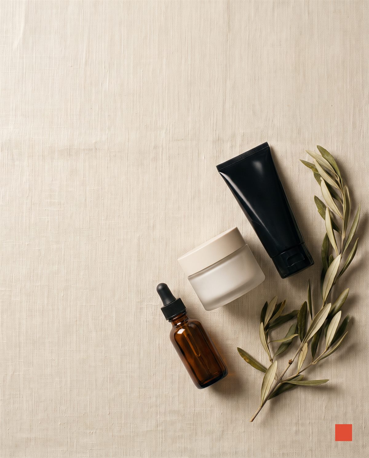 Flat lay of amber serum, cream jar and black tube on bone linen with olive leaves